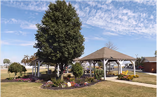 Outdoor area with two wooden gazebos surrounded by a garden with flowers and a large tree in the center. There is a paved walkway and a building visible in the background under a partly cloudy sky.