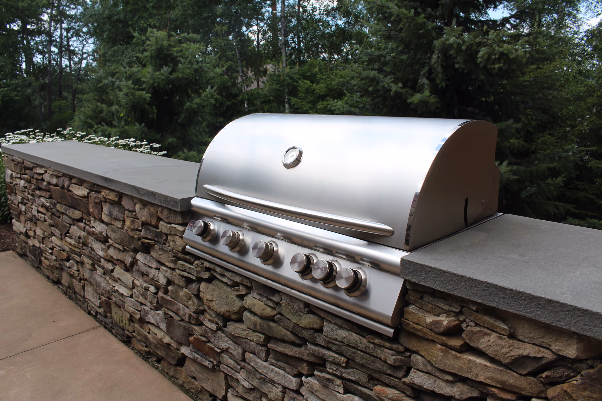 A stainless steel outdoor gas grill built into a stone countertop with a stone base, surrounded by greenery and trees in the background.