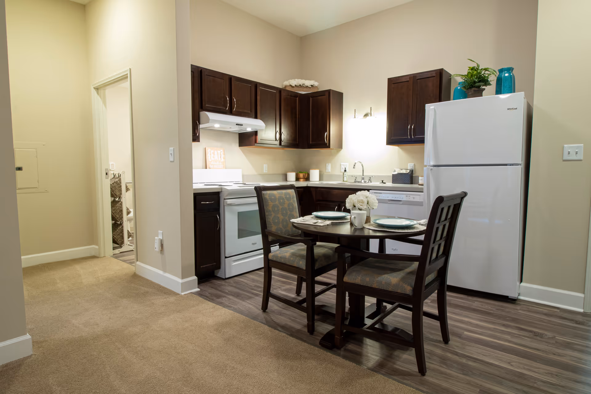 A small kitchen and dining area with dark wood cabinets, a white refrigerator, stove, and dishwasher. There is a round dining table set for two with plates, cups, and a small flower arrangement. The floor transitions from carpet in the hallway to wood laminate in the kitchen area. The walls are painted light beige.
