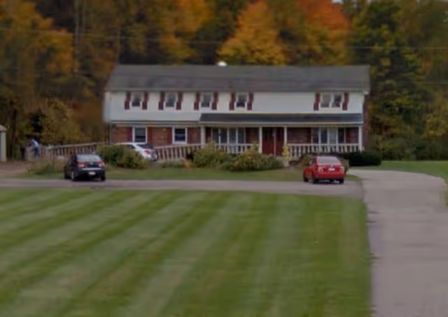 Front view of a two-story house with a wide porch, a large mowed lawn, a driveway, and two parked cars with autumn trees in the background.