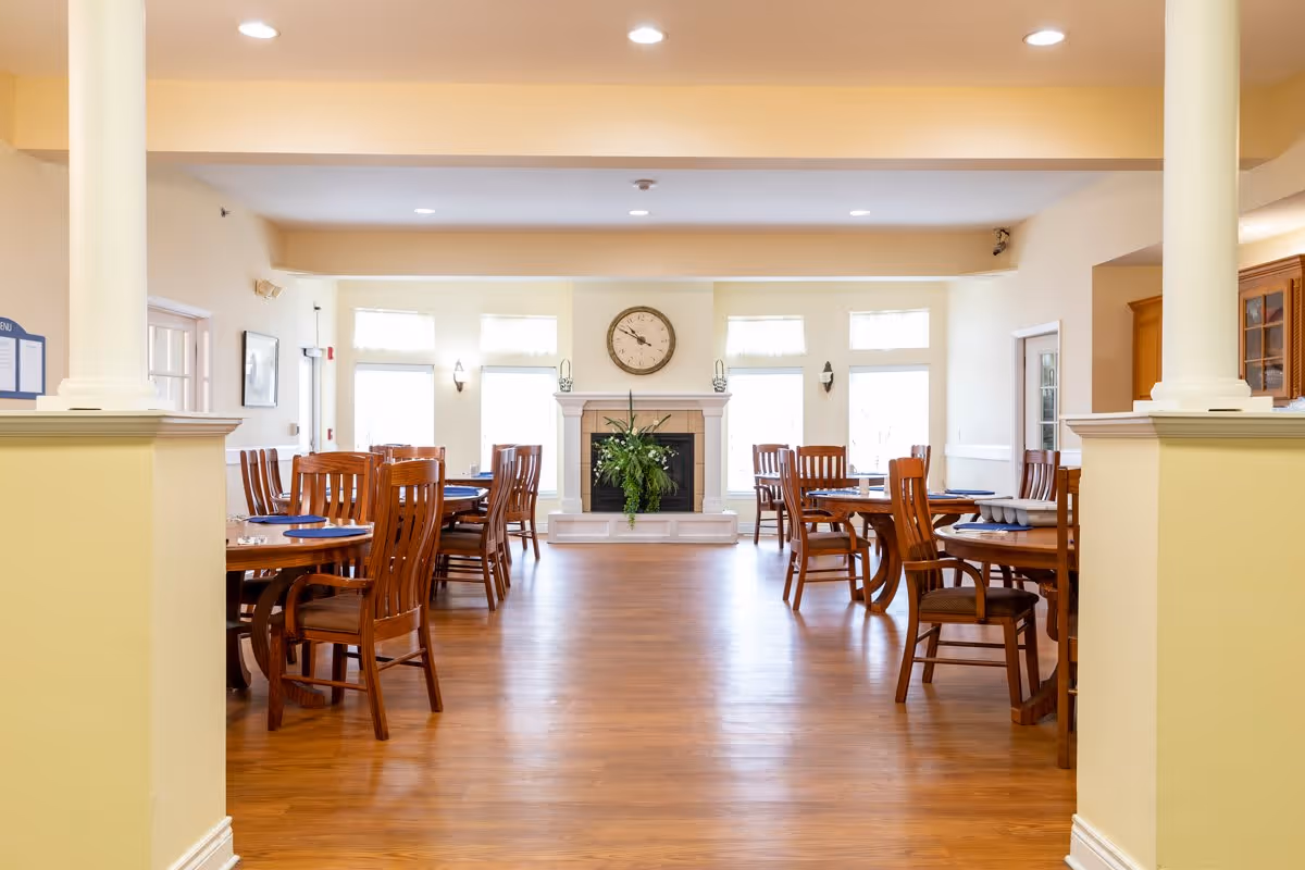 A bright and spacious dining room with wooden floors and several round wooden tables surrounded by wooden chairs. The room features large windows letting in natural light, a fireplace with a clock above it, and light-colored walls. The setting is neat and inviting, suitable for communal dining.