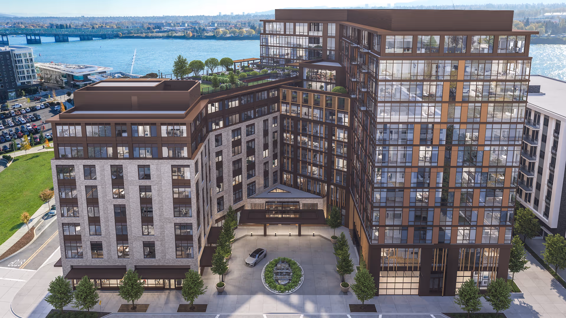 Aerial view of a modern multi-story residential building complex with large glass windows and a central courtyard featuring a circular planter with greenery. The building is situated near a waterfront with a river and bridge visible in the background. Trees and parked cars are seen around the building.
