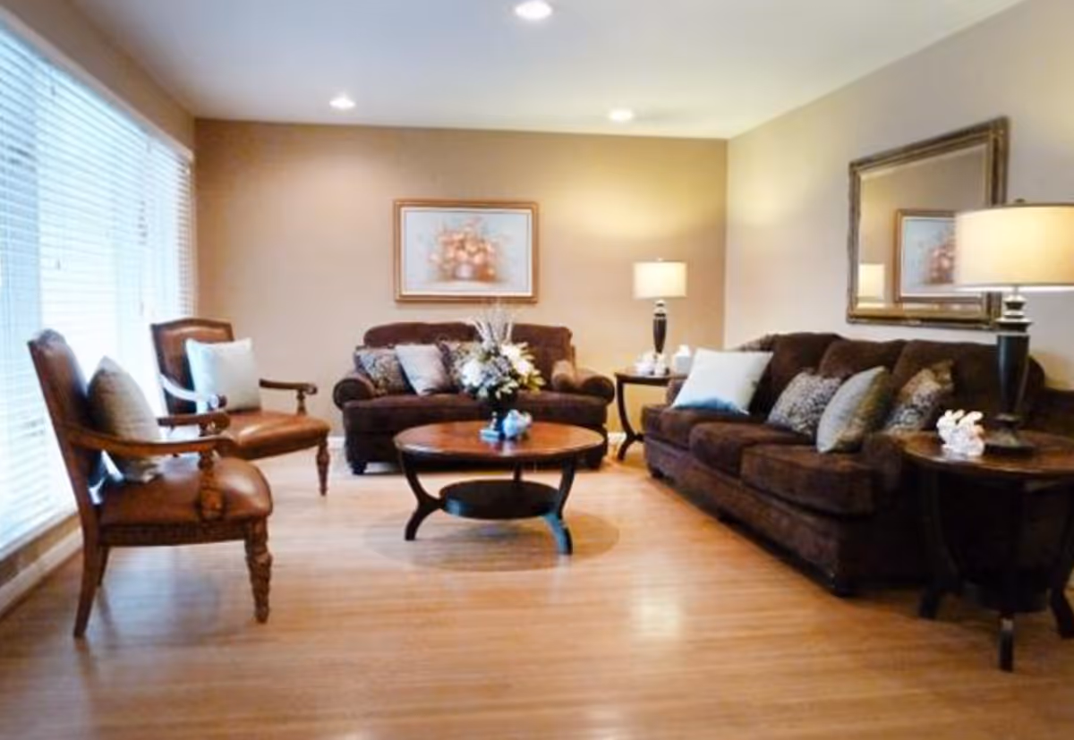 A cozy living room with two brown sofas adorned with multiple cushions, two wooden armchairs with cushions, a round wooden coffee table with a floral arrangement, two side tables each with a lamp, a large mirror on the wall, and a framed floral picture. The room has wooden flooring and large windows with blinds allowing natural light.