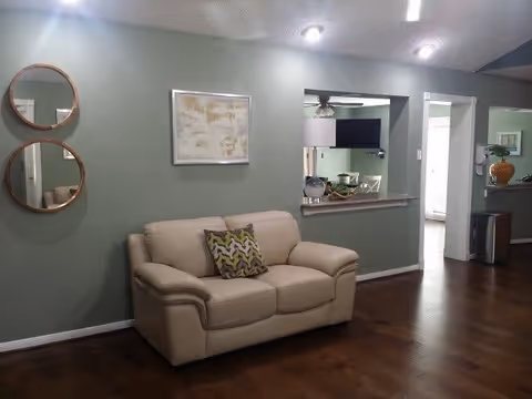 A cozy living room area with a beige loveseat featuring a patterned cushion. The wall behind the loveseat is painted green and decorated with two round mirrors and a framed artwork. There is a pass-through window to the kitchen area, where a TV and some kitchen items are visible. The floor is dark wood, and the room is softly lit by ceiling lights.