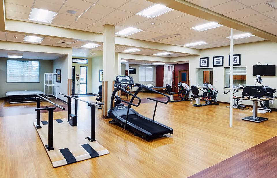 Well-lit rehabilitation gym room with a treadmill, parallel bars, exercise bikes and other fitness equipment on wood floors.