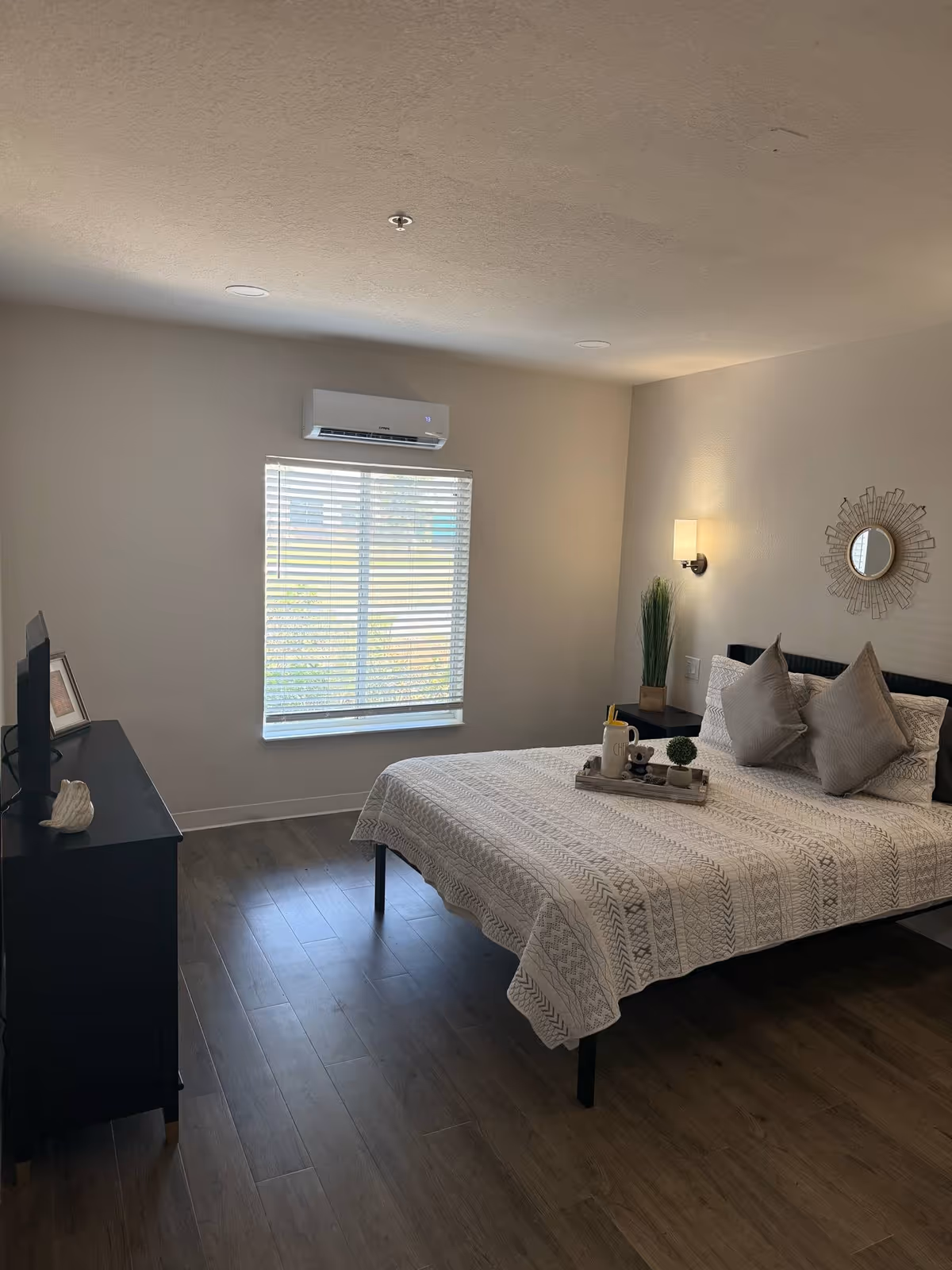 A neatly arranged bedroom with a bed covered in a patterned white and gray bedspread, two large pillows, and a tray holding a mug, a small plant, and a decorative item. The room has wooden flooring, a window with blinds, a wall-mounted air conditioner above the window, a black dresser with a TV and decorative items, a nightstand with a tall plant, a wall-mounted light, and a decorative round mirror on the wall.