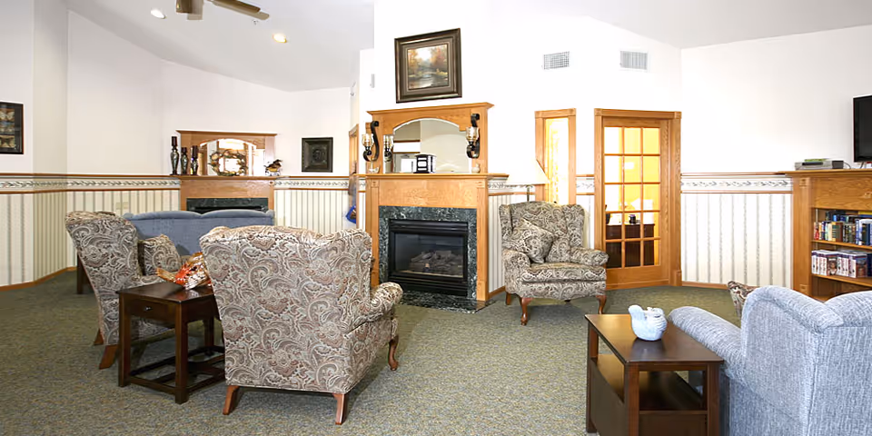 A cozy senior living facility common area with patterned armchairs, a gray sofa, wooden side tables, a fireplace with a mirror and sconces above it, a wooden bookshelf with books and DVDs, and a door with glass panels. The walls are white with a striped wallpaper border and the carpet is greenish.
