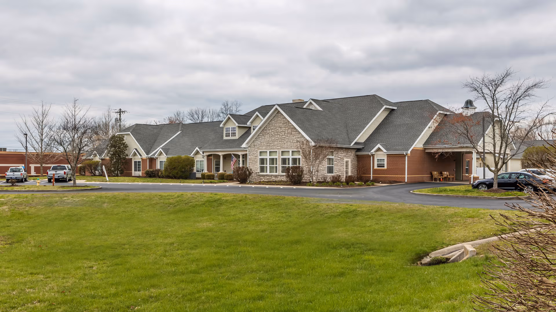 Exterior view of a single-story senior living facility building with a combination of brick and stone facade, multiple gabled roofs, and a driveway with parked cars. The foreground features a well-maintained green lawn and some leafless trees under a cloudy sky.