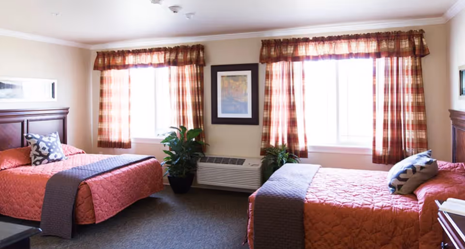 A bedroom with two beds covered in orange quilted bedspreads and gray throws. Each bed has a decorative pillow. There are two windows with plaid curtains letting in natural light. Between the windows is a framed picture on the wall and an air conditioning unit below it. Two potted plants are placed on either side of the air conditioner. The room has beige walls and carpeted flooring.