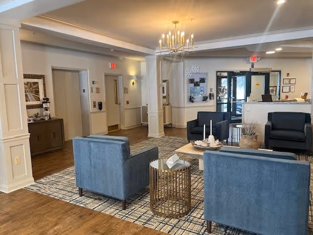 A spacious lobby area featuring blue armchairs, a decorative coffee table, and a reception desk in the background. The space is well-lit with a chandelier and has a warm, inviting atmosphere.