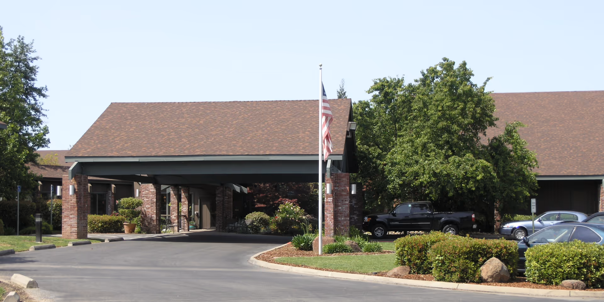 Covered entrance canopy of a senior living facility with a flagpole, parked cars, and landscaped driveway.