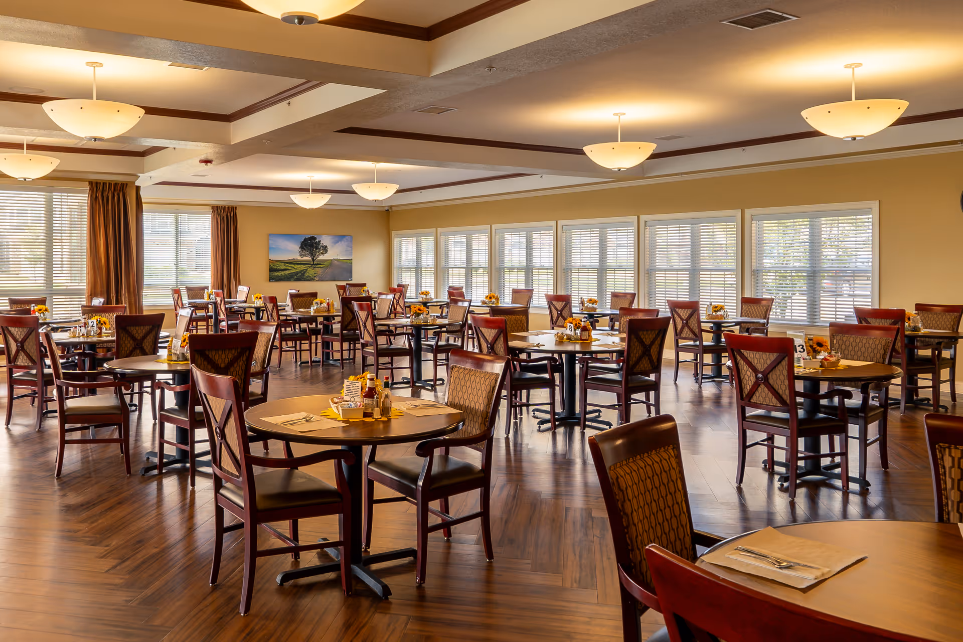 A spacious dining room with multiple round wooden tables and cushioned chairs arranged neatly. Each table is set with placemats, utensils, and condiments. Large windows with blinds allow natural light to fill the room, and a landscape painting hangs on the far wall. The floor has a wood finish, and ceiling lights provide warm illumination.