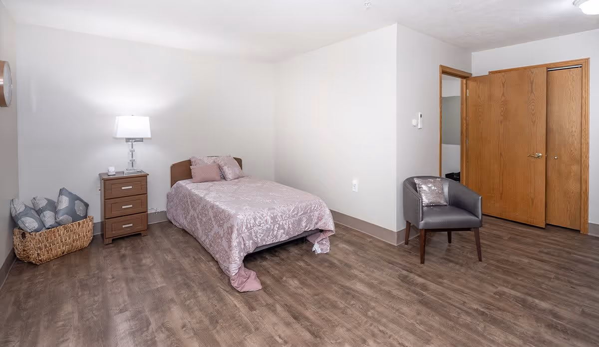 A simple bedroom with a single bed covered in a light purple patterned bedspread and matching pillows. Next to the bed is a wooden nightstand with a lamp and a small item on it. There is a basket with several pillows on the floor to the left. A gray armchair with a shiny pillow is positioned near a wooden closet with double doors. The room has wood flooring and plain white walls.
