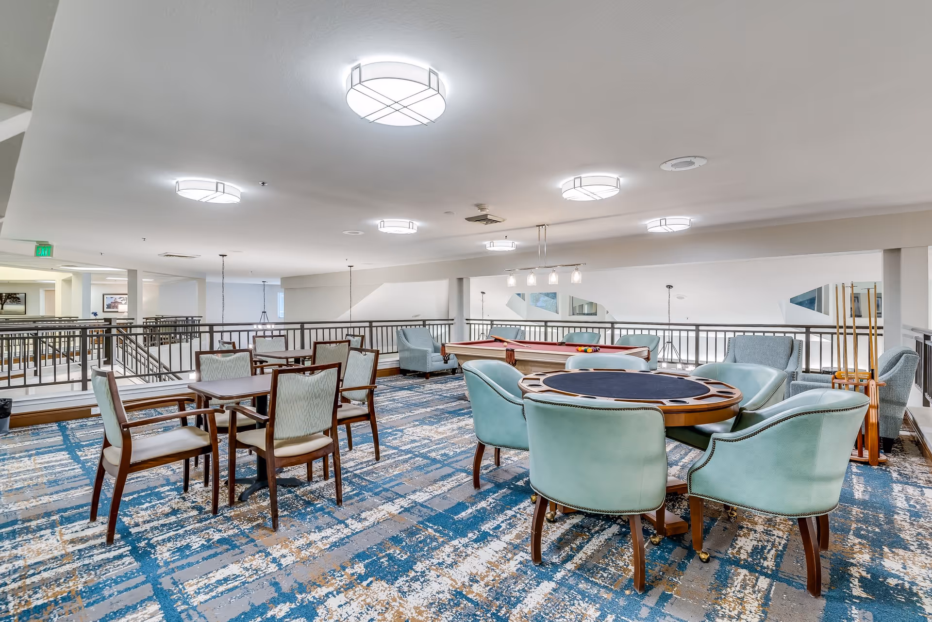 A spacious senior living common area featuring a card table with six teal upholstered chairs, a pool table with cues, and several wooden tables with chairs. The room has modern ceiling lights, a patterned carpet in blue and beige tones, and a railing overlooking a lower level.