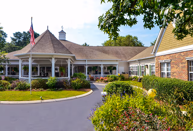 Exterior view of a senior living facility with a circular driveway, landscaped garden beds, and an American flag on a flagpole. The building features a covered porch with white railings and a combination of brick and siding on the walls. Trees and shrubs surround the area under a partly cloudy sky.