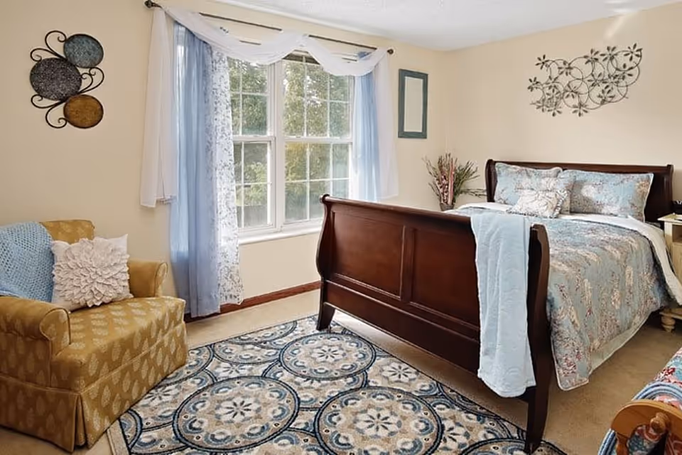 Bright furnished bedroom with a wooden sleigh bed, patterned bedding, an armchair, large window and decorative rug.