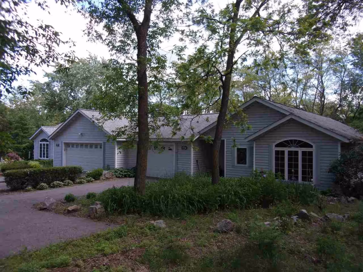 Exterior view of a single-story residential building surrounded by trees and greenery. The building has light gray siding, multiple garage doors, and large windows. A paved driveway leads up to the garages, and there are bushes and plants around the property.