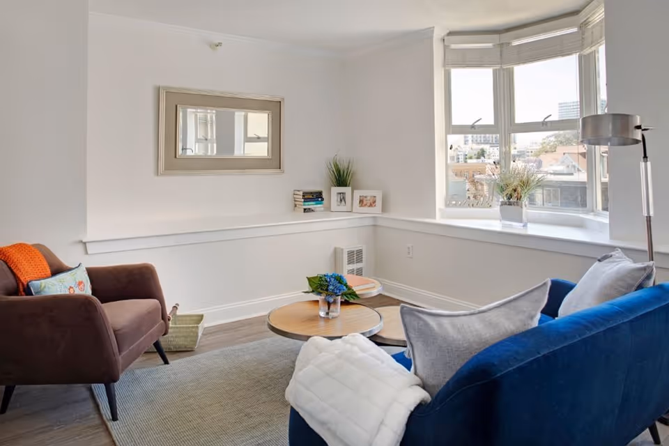 A cozy living room with a blue sofa adorned with gray pillows and a white throw blanket, a brown armchair with a colorful pillow and orange throw, a round wooden coffee table with a small plant and books, a large window with a view of buildings outside, a floor lamp, and a wall mirror above a white ledge holding books and framed photos.