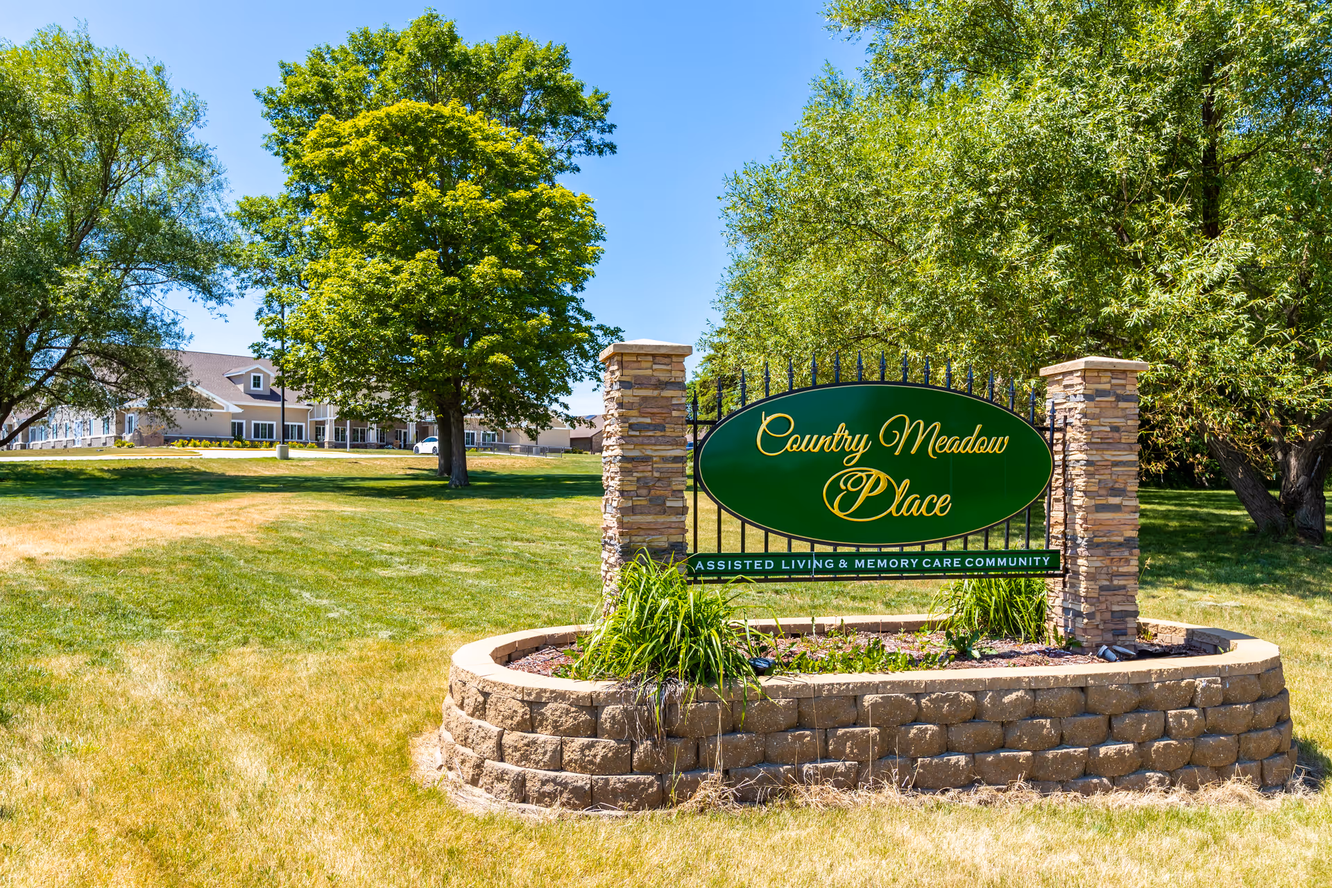 A large green and yellow sign reading 'Country Meadow Place Assisted Living & Memory Care Community' is displayed on a stone base with two stone pillars on a grassy lawn. Behind the sign, there are large trees and a building with a brown roof and white trim under a clear blue sky.