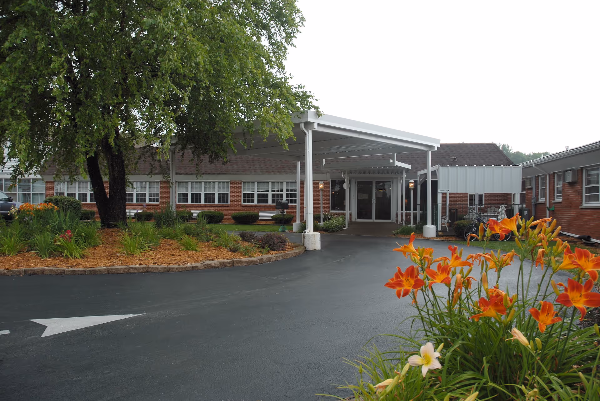 Covered entry and drop-off drive of a one-story brick care facility with landscaping and orange flowers in the foreground.