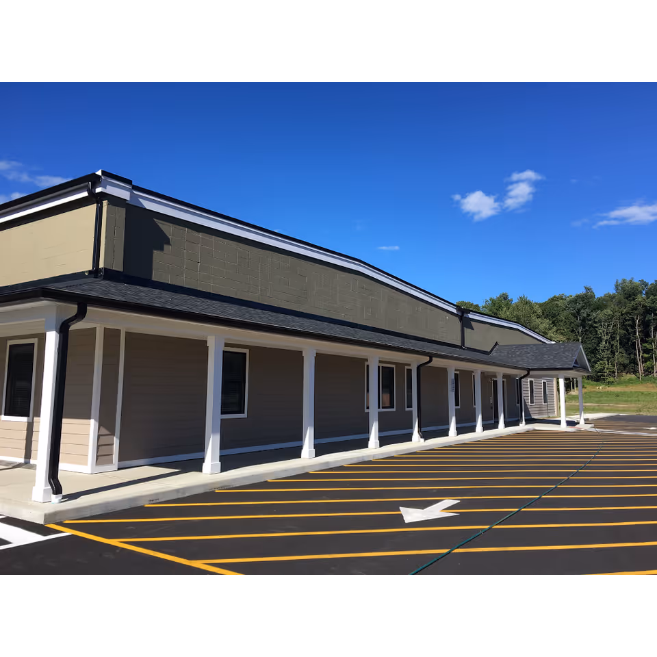 Exterior view of a single-story building with beige siding and white columns supporting a covered walkway. The building is adjacent to a parking lot with freshly painted yellow lines and a white directional arrow. The sky is clear and blue with a few clouds, and there are trees in the background.