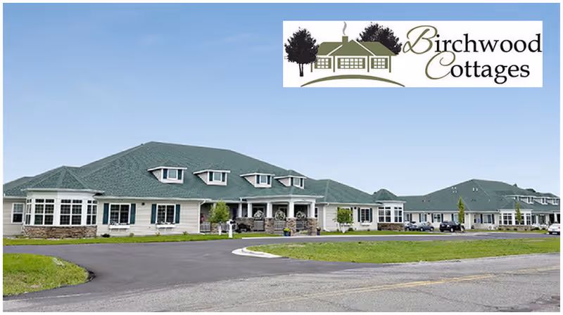 Exterior view of a single-story senior living facility with green roofs and multiple windows, surrounded by a paved driveway and green lawn under a clear blue sky. The facility is named Birchwood Cottages, as indicated by the logo in the top right corner.