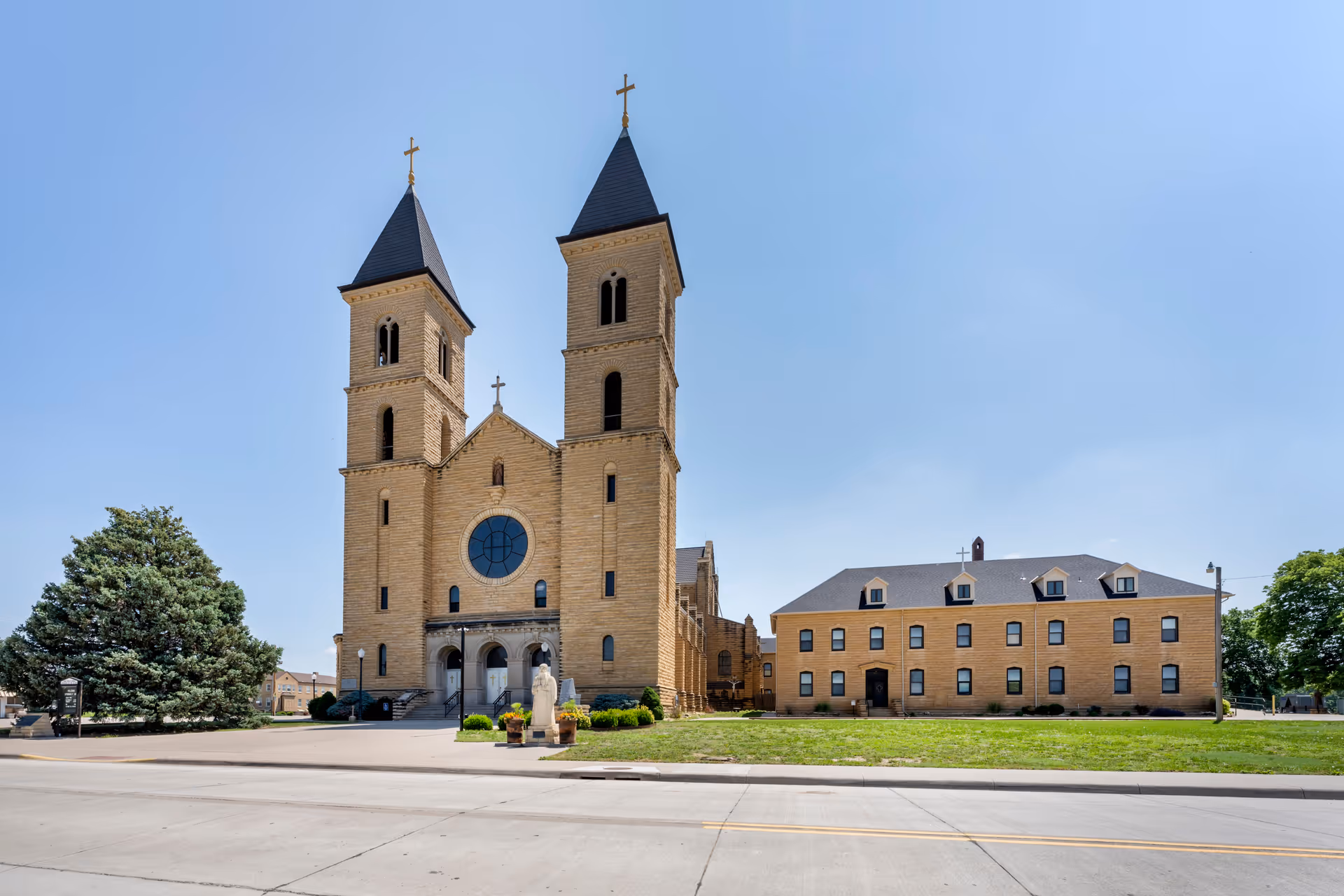 Exterior view of a large stone church with two tall towers topped with crosses, adjacent to a three-story brick building with multiple windows and dormer windows on the roof, under a clear blue sky.