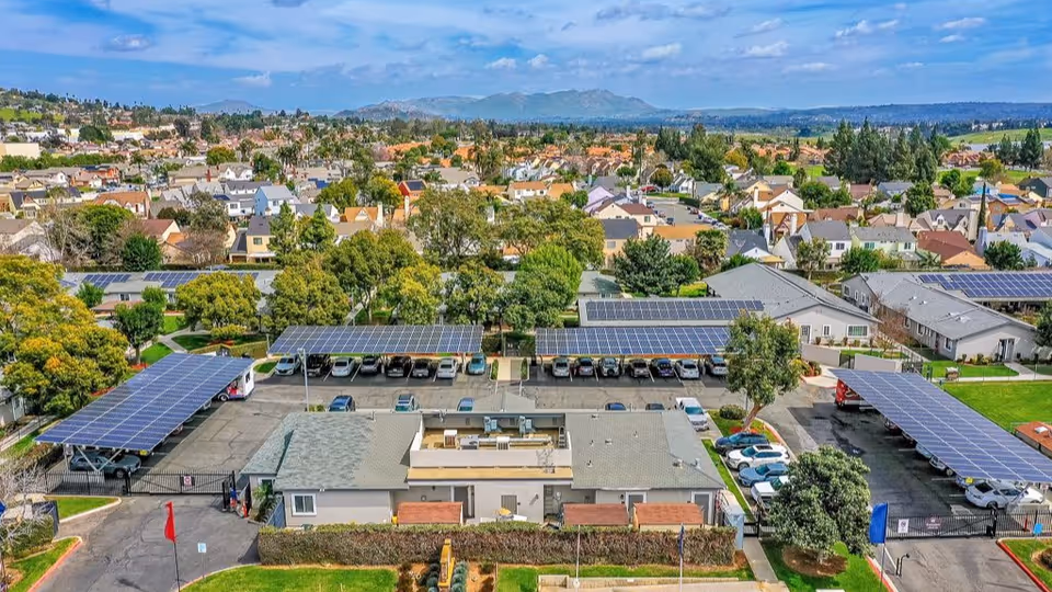 Aerial view of a senior living facility named Cottages at Riverside, showing multiple single-story buildings with solar panel-covered parking areas, surrounded by trees and residential neighborhoods under a partly cloudy sky.