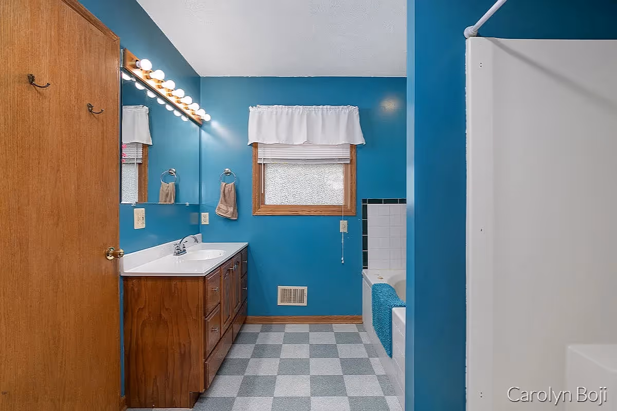 A bathroom with blue walls and a checkered gray and white floor. There is a wooden vanity with a white countertop and a sink, a large mirror with lights above it, a window with a white valance, a towel hanging on a ring, and a bathtub with a blue towel draped over the edge. A wooden door is partially visible on the left side.
