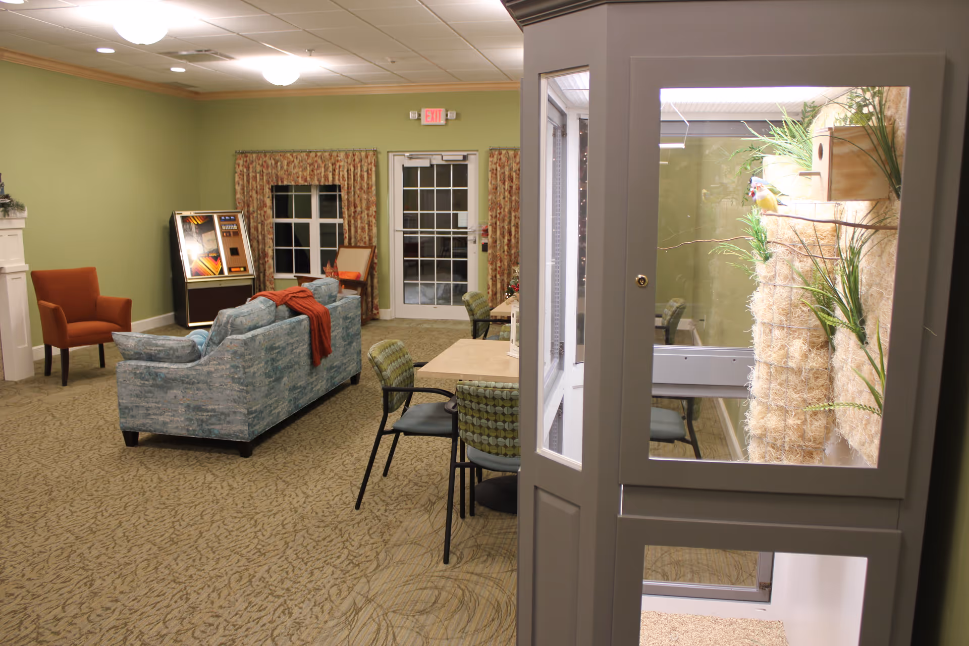 A cozy living room area in a senior living facility with green walls and patterned carpet. The room features a blue patterned sofa with a red throw blanket, an orange armchair, a small table with chairs, and a jukebox in the corner. There is a glass-enclosed bird habitat with plants and a bird perched inside on the right side of the image. A door and window with floral curtains are visible in the background.
