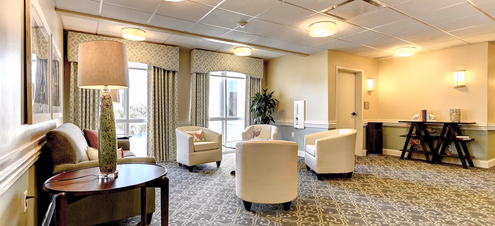 A bright and spacious living room area in a senior living facility with patterned carpet, four white armchairs arranged around a small table, a sofa with cushions, a tall table lamp, large windows with patterned curtains, and a decorative console table with books and ornaments against the wall.