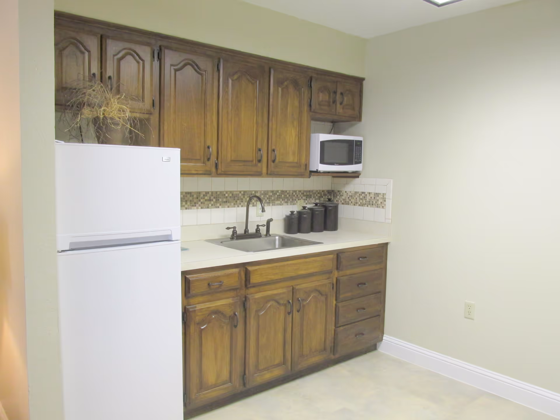 A small kitchen area with wooden cabinets, a white refrigerator, a microwave mounted above the counter, a sink with a faucet, and several black canisters on the countertop. The backsplash features a mosaic tile design.