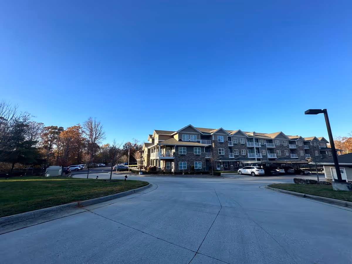 Exterior view of a three-story senior living building with parked cars, a driveway, and a clear blue sky.