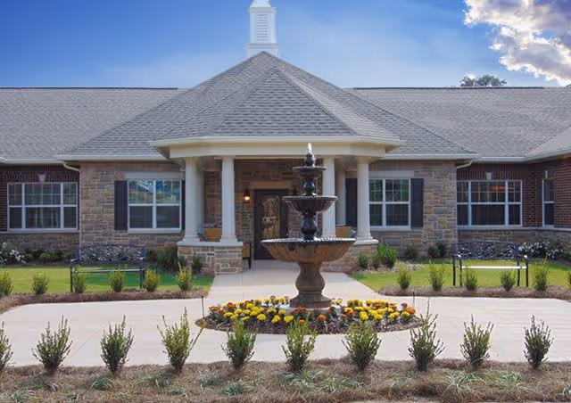 Front exterior view of a single-story brick and stone building with a gray shingled roof, white columns at the entrance, a three-tiered black fountain surrounded by flowers in the foreground, and a clear blue sky with some clouds above.