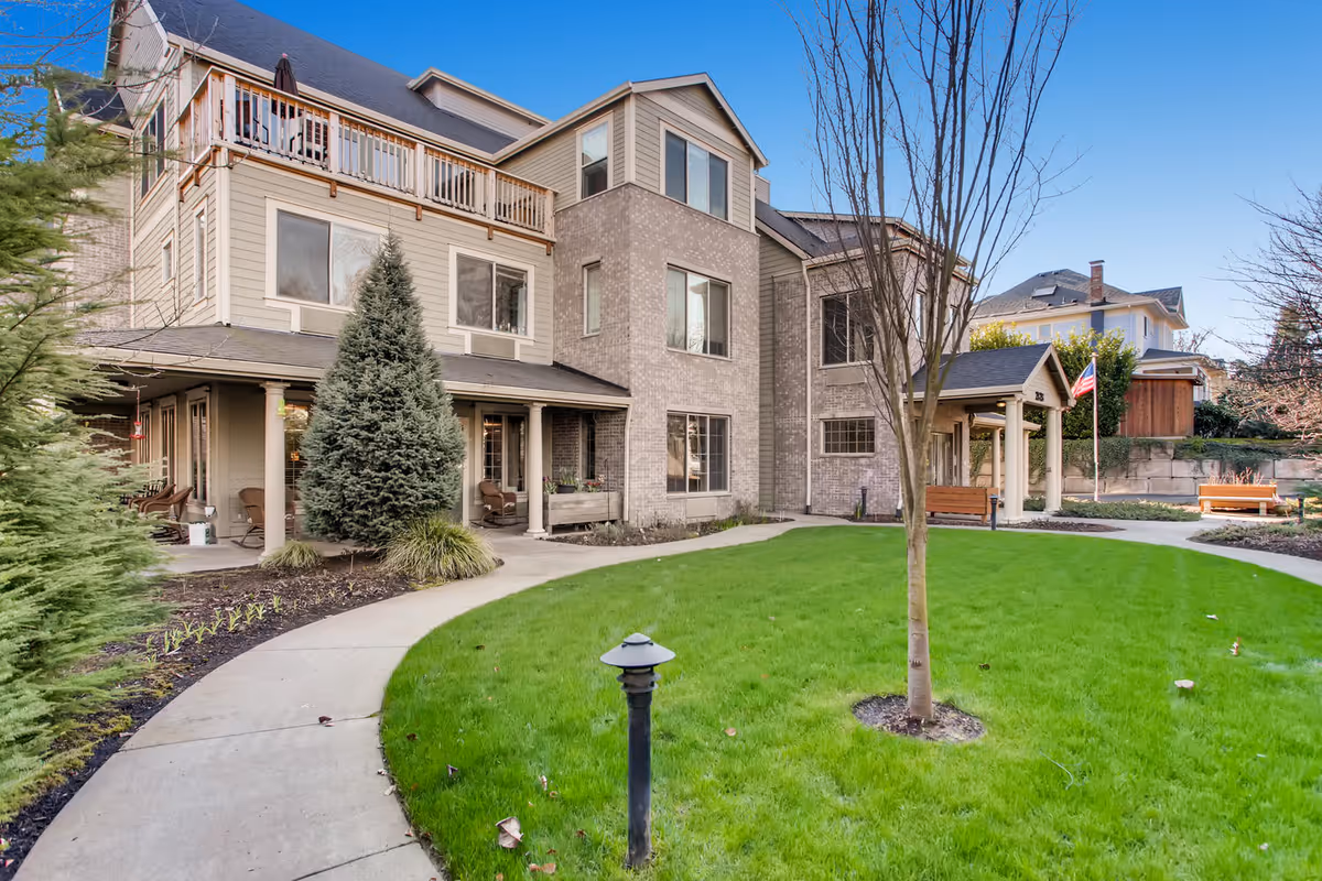 Exterior view of a senior living community building with a well-maintained lawn, a curved concrete pathway, several trees, and benches. The building has multiple floors with large windows, a balcony, and a covered entrance. The sky is clear and blue.