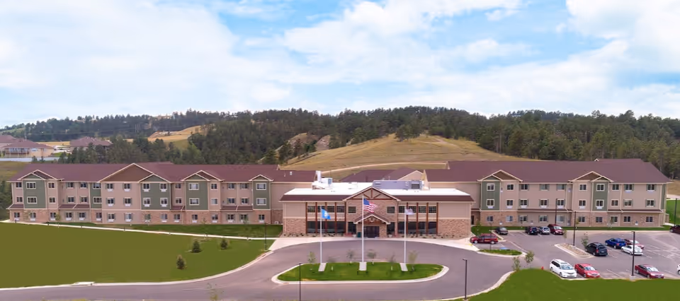 Front exterior of a long three-story assisted living building with a circular drive, flagpoles, parking lot, and wooded hills behind it.