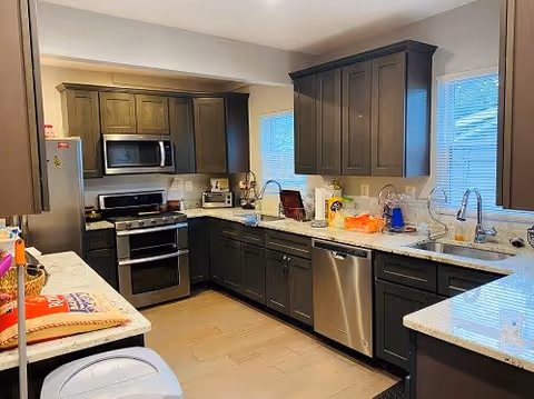 Modern kitchen with dark wood cabinets, stainless steel appliances including a refrigerator, microwave, oven, and dishwasher. The kitchen has light-colored countertops and two windows with blinds letting in natural light. Various kitchen items and small appliances are placed on the countertops.