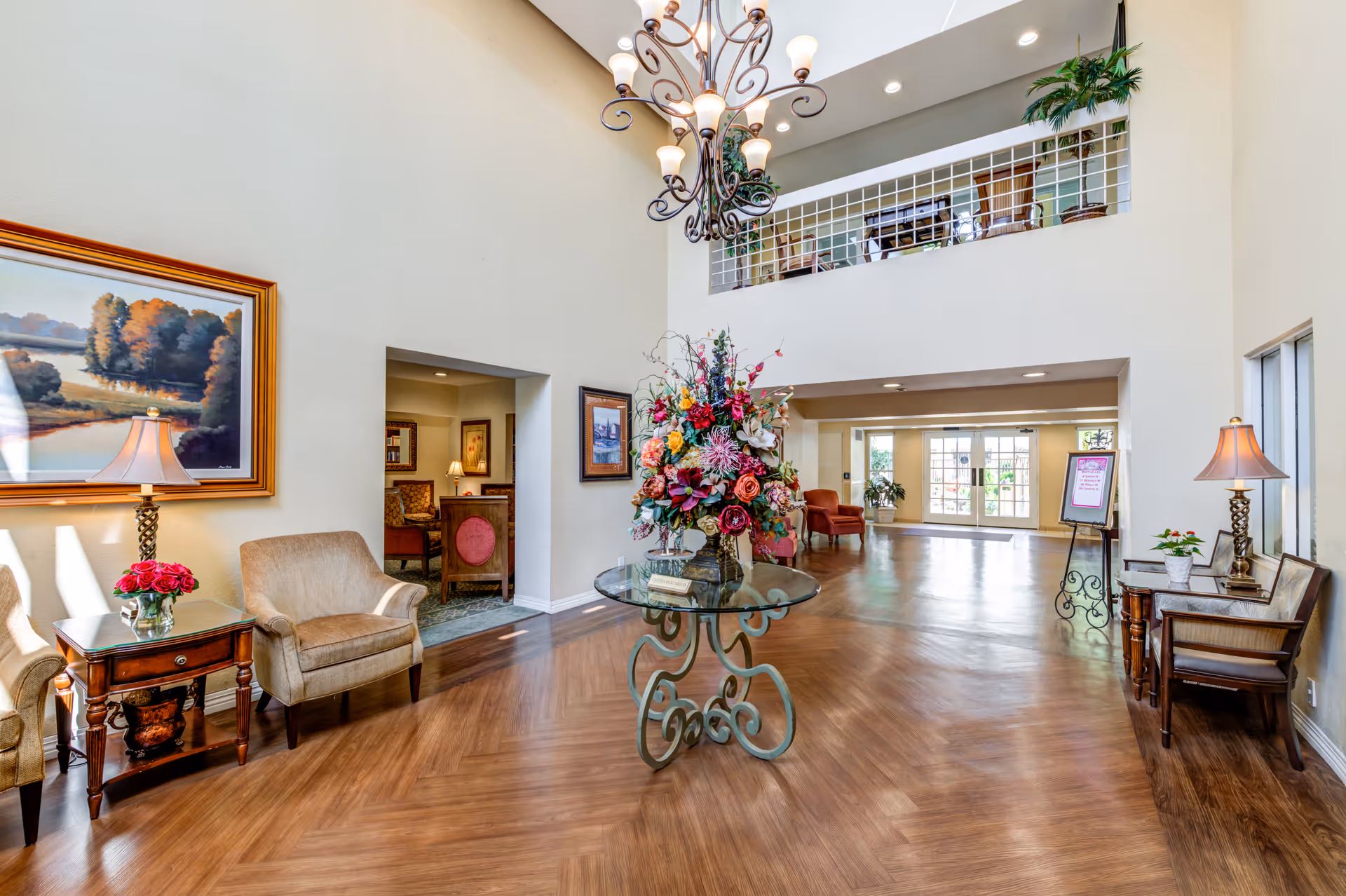 A spacious and well-lit interior lobby area with high ceilings and wooden flooring. In the center, there is a round glass table with an ornate metal base holding a large colorful floral arrangement. On the left side, there are two beige armchairs with a wooden side table between them, topped with a lamp and a small vase of red flowers. The walls are decorated with framed landscape paintings. On the right side, there is a wooden bench with a lamp and a small plant on a side table. A chandelier hangs from the ceiling, and in the background, there is a seating area with chairs and large windows letting in natural light.