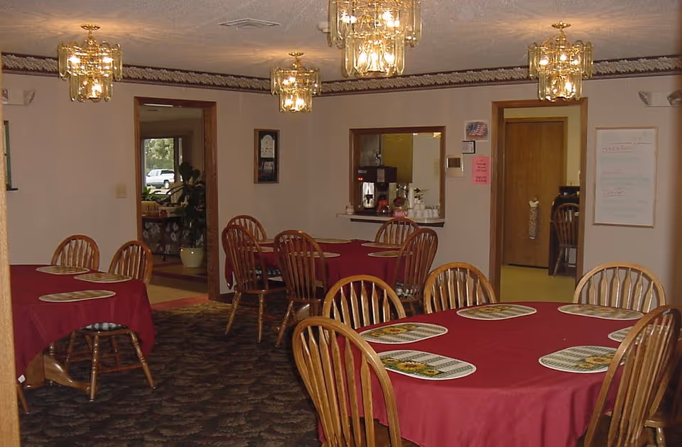 Dining room with multiple round tables covered with red tablecloths and wooden chairs around them. Each table has placemats with sunflower designs. The room has patterned carpet, beige walls, and several glass chandeliers hanging from the ceiling. There are doorways leading to other rooms, one with a coffee station visible through a pass-through window.