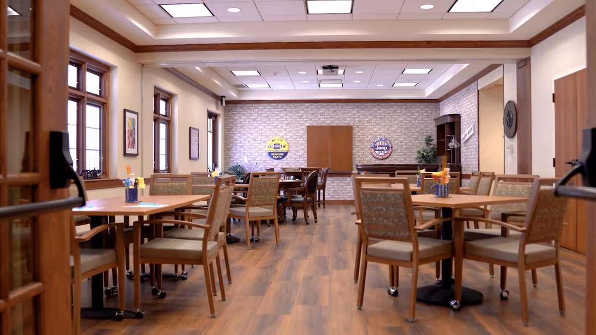 A spacious dining room with wooden floors and multiple tables surrounded by chairs. The room has large windows on the left wall, framed artwork, and a brick accent wall at the far end with vintage automotive signs. The ceiling has recessed lighting and a projector mounted in the center.