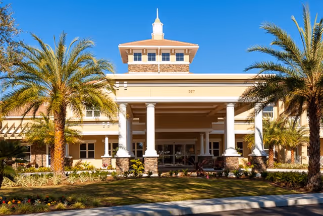Front exterior view of a senior living facility building with a covered entrance supported by white columns, palm trees, and landscaped greenery under a clear blue sky.