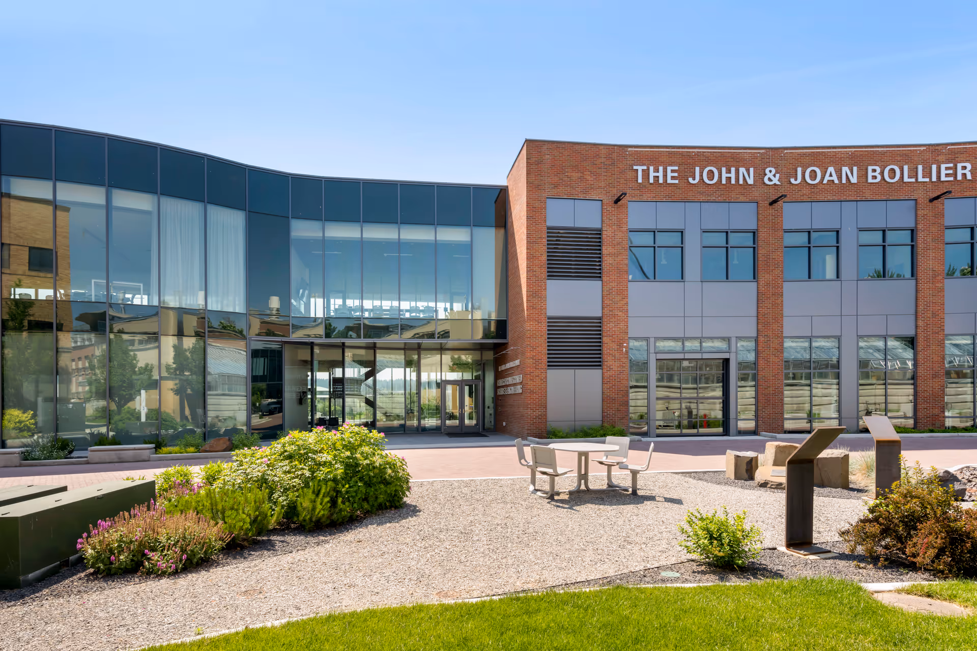 Exterior view of a modern building with large glass windows and brick walls under a clear blue sky. In front of the building, there is a landscaped area with green bushes, gravel, a round table with four chairs, and some stone benches.