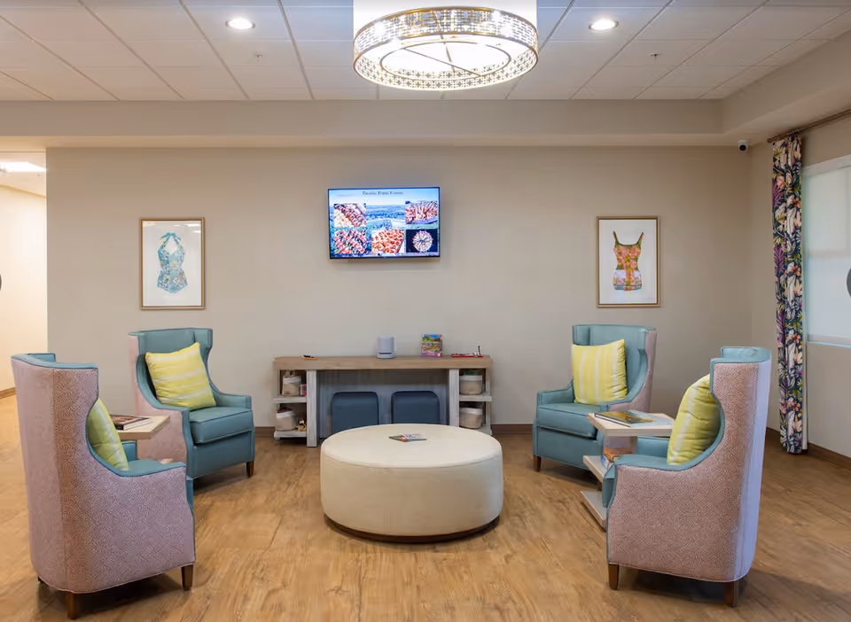 Communal lounge with four pastel upholstered armchairs around a round ottoman, a console table and a wall-mounted TV.