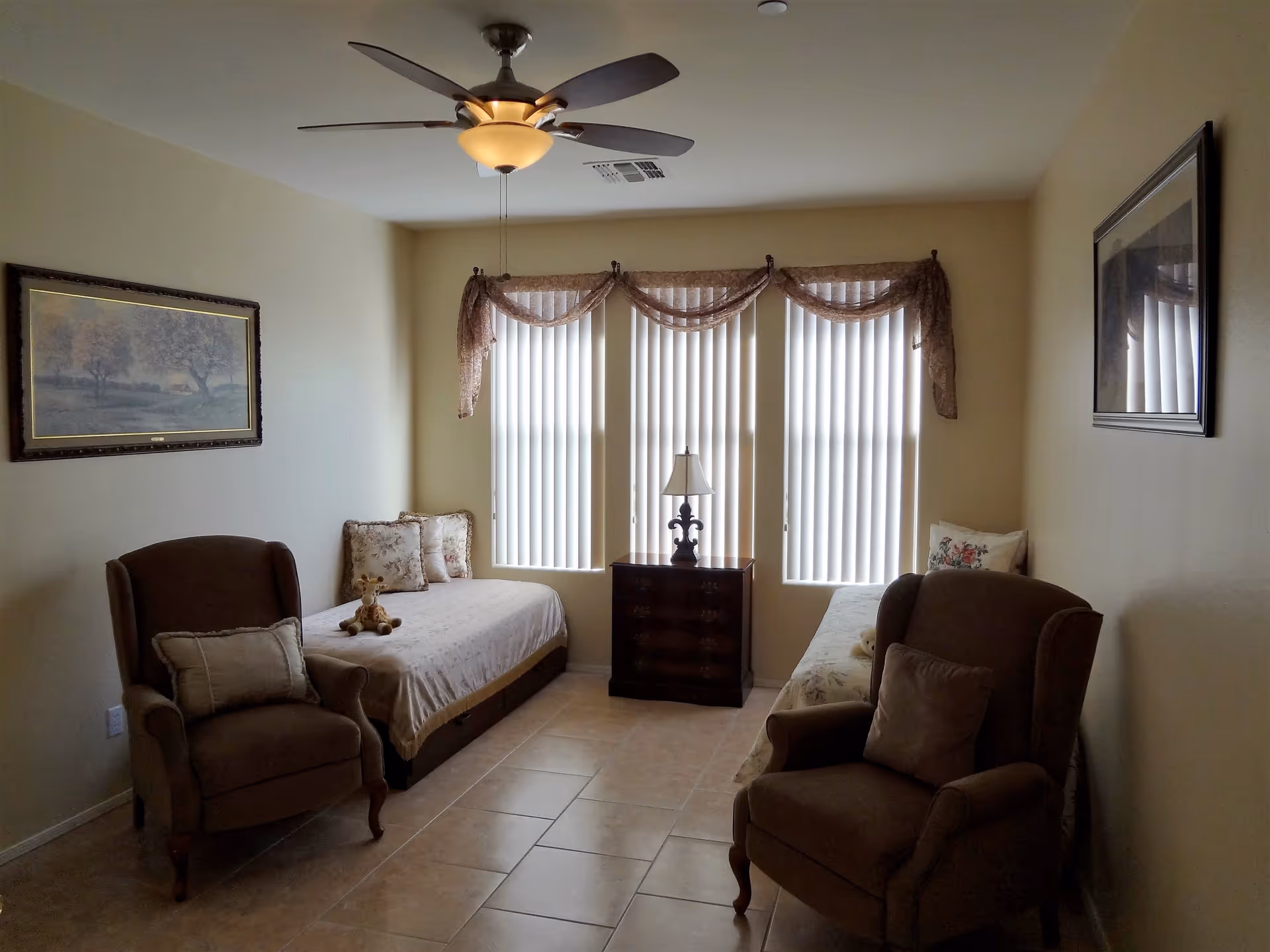 Small living room with two armchairs, two daybeds, a central side table with a lamp, and three tall windows with vertical blinds.