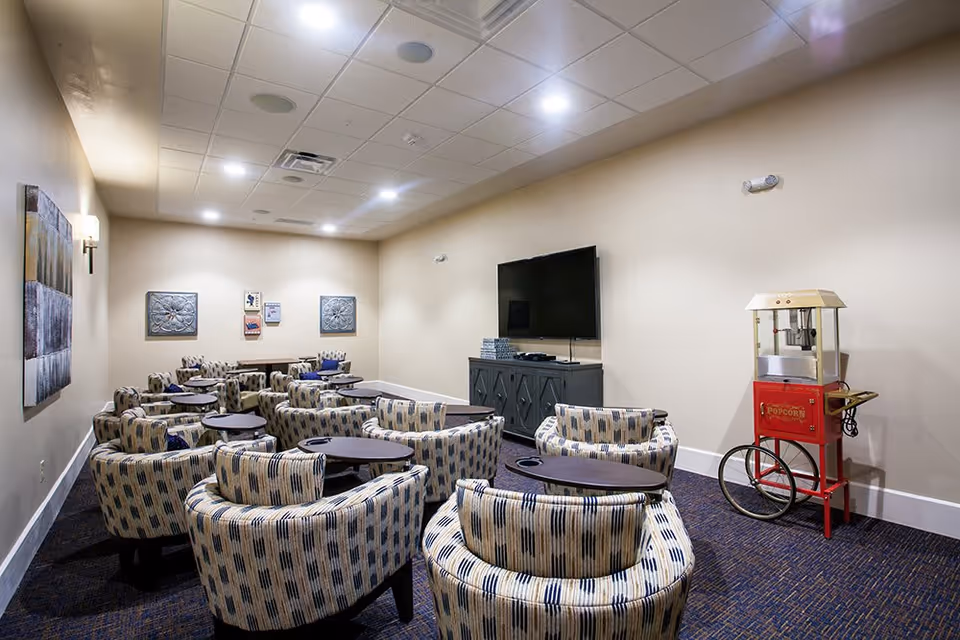 A cozy media room with multiple patterned armchairs arranged in rows facing a large flat-screen TV mounted on a cabinet. There is a vintage-style red popcorn machine on wheels to the right side of the room. The walls are beige with some decorative wall art, and the ceiling has recessed lighting.