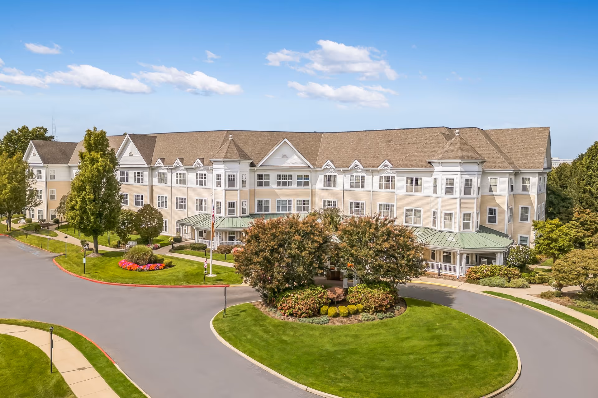 Exterior view of a large, three-story senior living facility building with beige siding and white trim, surrounded by well-maintained landscaping including green lawns, trees, and flower beds under a blue sky with scattered clouds.