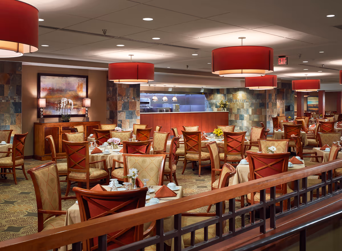 A spacious dining room with multiple tables covered in beige tablecloths, each set with white dishes, glassware, and folded red napkins. The chairs have wooden frames with red and beige upholstery. The room features large red pendant lights hanging from the ceiling, stone accent walls, and a sideboard with a framed abstract painting and two lamps. The kitchen area is visible in the background.