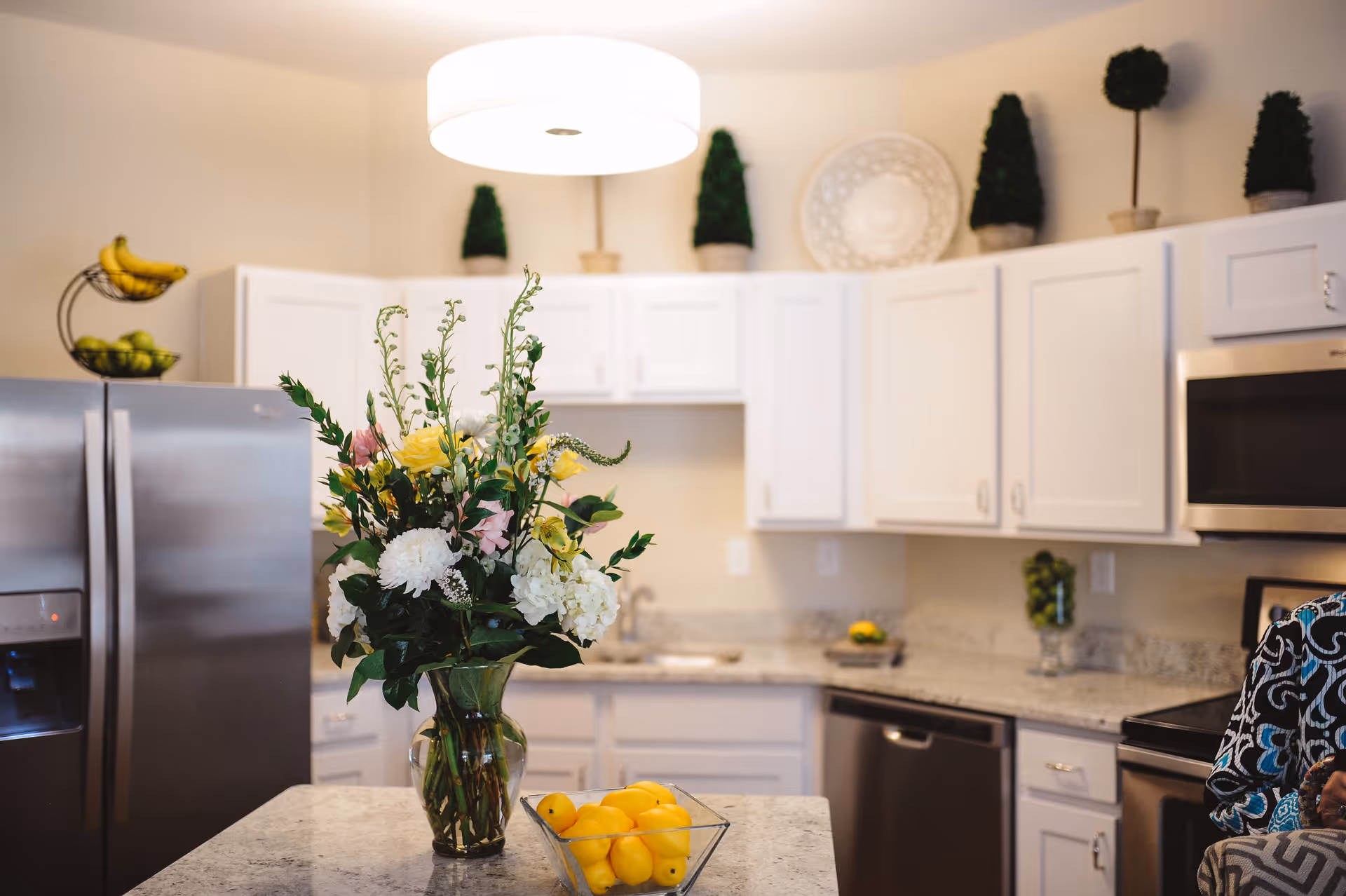 A modern kitchen with white cabinets, stainless steel appliances including a refrigerator and microwave, and a granite countertop island. On the island, there is a vase with a colorful flower arrangement and a glass bowl filled with lemons. Decorative topiary plants and a large decorative plate are displayed on top of the cabinets. A bright circular ceiling light illuminates the space.