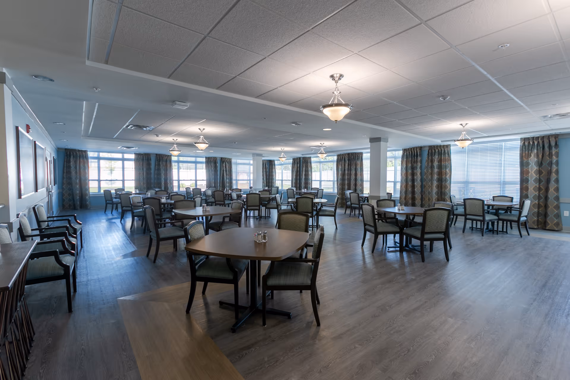 A spacious dining room with multiple round tables and chairs arranged neatly. The room has large windows with patterned curtains allowing natural light to fill the space. The ceiling has several light fixtures, and the floor is covered with wood-like flooring.