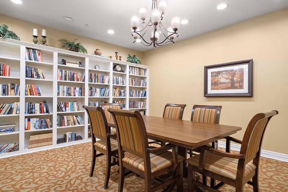 A well-lit room with a wooden table surrounded by six striped upholstered chairs. Behind the table is a large white bookshelf filled with books and decorative items. The walls are painted beige, and there is a framed picture of a tree on the wall. A chandelier with multiple lights hangs from the ceiling, and the floor is covered with a patterned carpet.